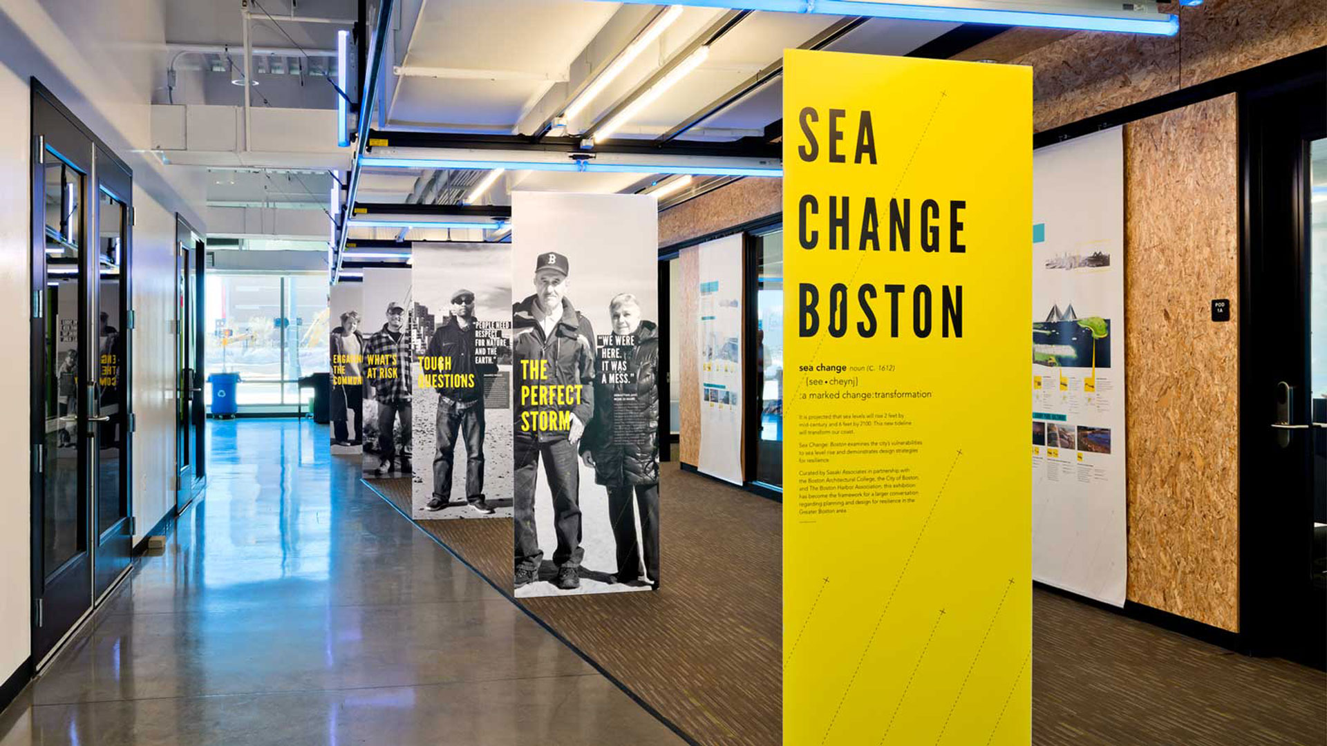 A photograph of a hallway with blue lighting and a series of 5 pillars in line in the hallway. The pillar in the front is yellow with typograhpy. The following pillars are all photographs of people in black and white with yellow typography. In the background there are plywood walls with large posters hanging on them. Design made in Adobe Indesign, Adobe Illustrator, and Adobe Photoshop.